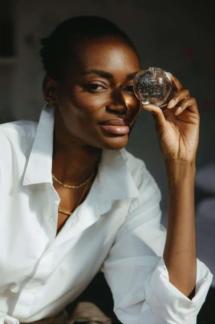 Portrait of an African American woman in a white collared shirt holding a glass orb in front of her eye, showcasing a bright indoor scene.
