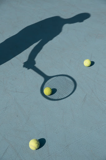 Overhead shot on sunny day shows shadow of tennis player with racket, and three yellow tennis balls on light blue court surface.