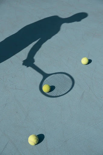 Overhead shot on sunny day shows shadow of tennis player with racket, and three yellow tennis balls on light blue court surface.