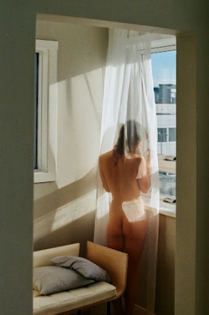 Rear view of a nude woman standing by a window partly veiled by sheer white curtains, photographed by Emily Winiker.