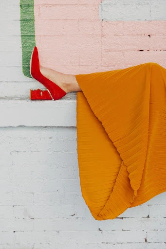 Woman's legs and red heels, seated on steps in front of colorful painted brick wall. Bright orange pleated skirt on figure.