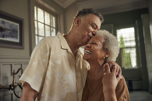 An older African-American couple embraces indoors; the man kisses the smiling woman on her forehead in a warm, tender moment.