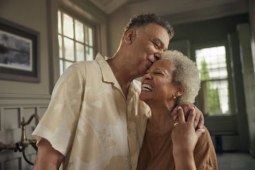 An older African-American couple embraces indoors; the man kisses the smiling woman on her forehead in a warm, tender moment.