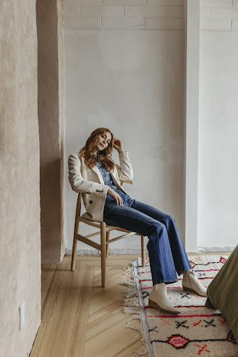 Red-haired woman in a cream blazer and jeans sits in a chair in a naturally lit room with light walls and a patterned rug.