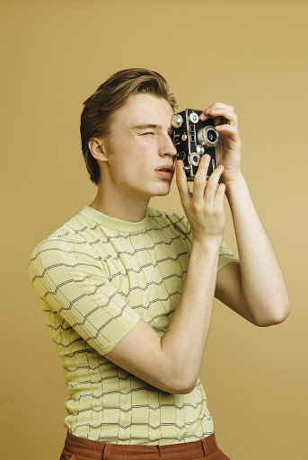 Young adult photographs with a vintage camera against a neutral background, highlighting retro style, photography, and a posed subject.