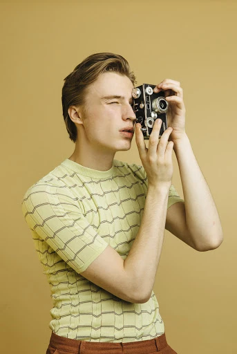 Young adult photographs with a vintage camera against a neutral background, highlighting retro style, photography, and a posed subject.