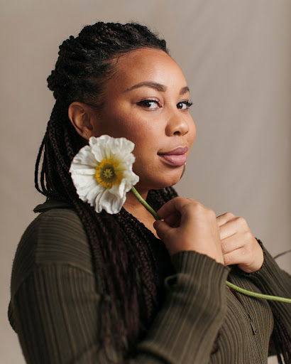 Portrait of a Black woman holding a white flower with a yellow center, wearing a dark green top against a neutral backdrop.