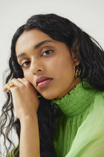 Close-up portrait of a young adult female with long dark hair wearing a green chiffon top and gold jewelry against a white background.