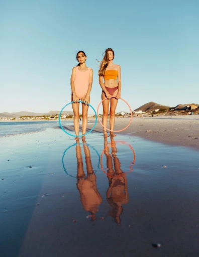 Two women in swimwear holding hula hoops stand at the ocean's edge on a sunny day, creating reflections in the wet sand.