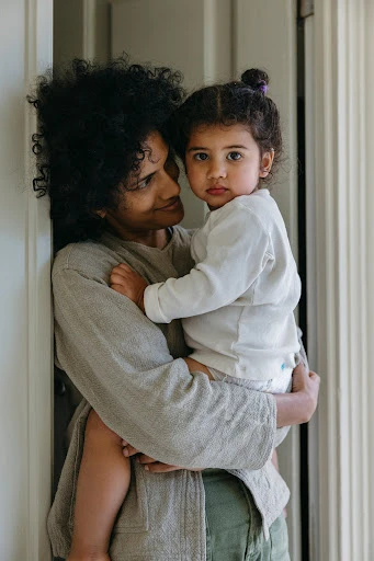 Indoor portrait of an adult woman with dark curly hair holding a young child with a serious expression in a neutral-toned room.