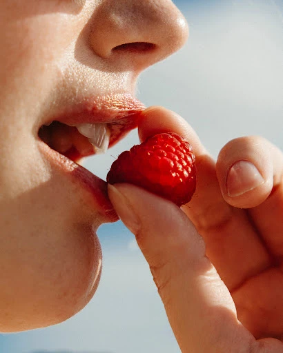 Close-up of a person eating a red raspberry held by their fingers against a light blue sky, highlighting the berry and the person's mouth.
