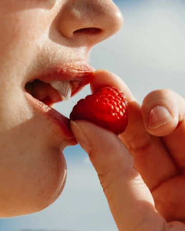 Close-up of a person eating a red raspberry held by their fingers against a light blue sky, highlighting the berry and the person's mouth.