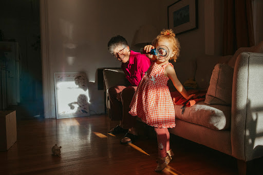 A young girl with red hair stands indoors holding a magnifying glass to her eye, with a woman seated on a sofa behind her.