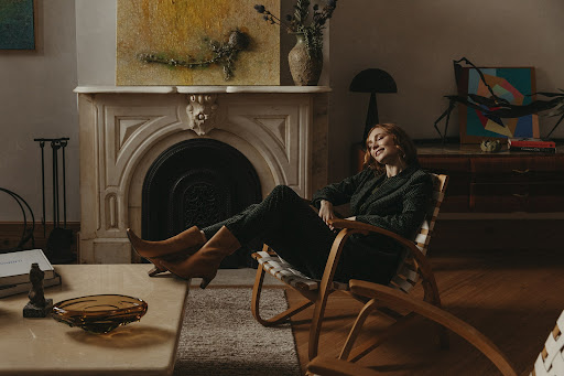 Red-haired woman relaxes in a wooden chair in a bright, vintage-decorated living room. Styled with tan boots and a patterned suit.