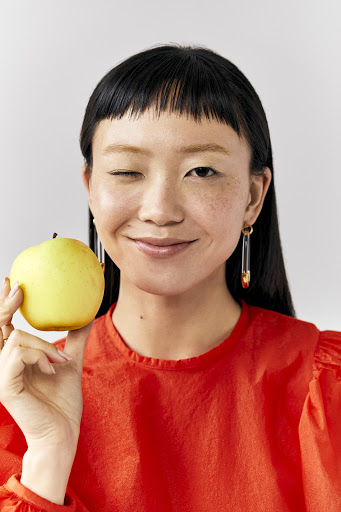 Attractive Asian woman winking and smiling while holding a bright green apple. She is wearing a red blouse with safety pin earrings.