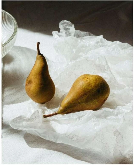 Two pears, one upright and the other lying on its side, are displayed on crumpled white paper, with a portion of a clear bowl visible.