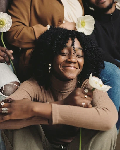 Smiling Black woman holds a white flower, surrounded by a diverse group in a warm studio setting, showcasing natural beauty and fashion.