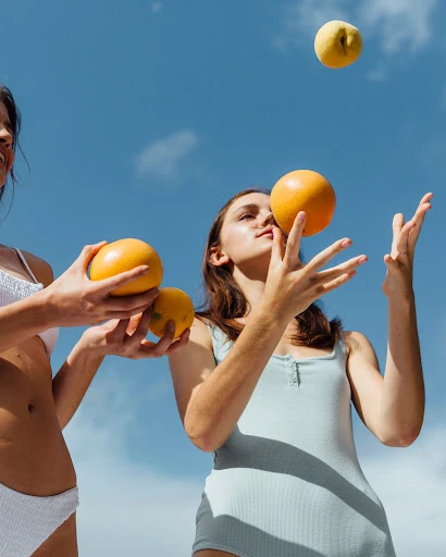 Two women in swimwear playing with fruit outdoors under a bright blue sky. One woman is juggling a grapefruit.