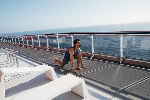 An adult male in fitness attire stretches barefoot on a cruise ship deck, featuring white lounge chairs, ocean view, and sunny sky.