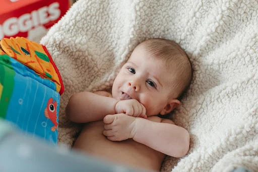 Infant lies on a textured blanket next to a colorful cube toy with an illustration of an orange fish and other aquatic animals.