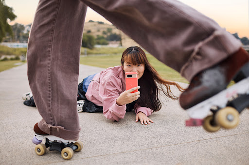 Two young women outdoors, one on roller skates with mauve jeans, another lying on ground with a cell phone, park setting.