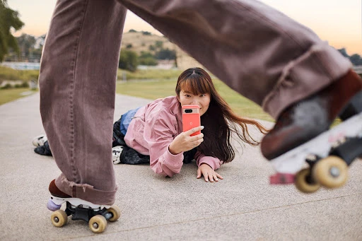 Two young women outdoors, one on roller skates with mauve jeans, another lying on ground with a cell phone, park setting.