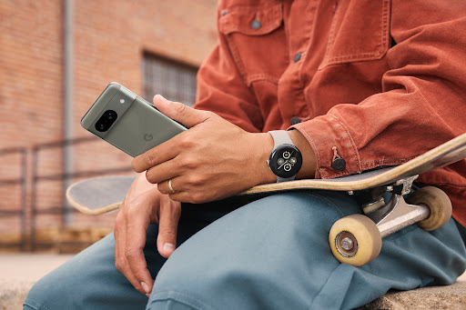 Person sitting with a skateboard, wearing a smart watch, and holding a mobile phone in an outdoor setting against a brick wall.