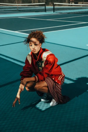 Young woman in a red varsity jacket and striped skirt crouching on a blue tennis court outdoors.