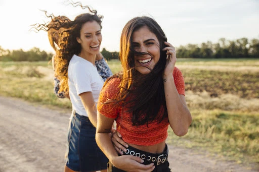 Three women laugh on a dirt road with golden hour sunlight, trees in the background and a field to the right. Joyful, friendship, fun.