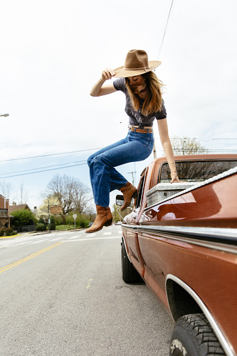 Woman in hat and jeans perches playfully on the truck bed's edge, mid-air, set against a bright daytime suburban street.