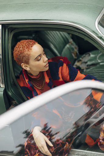 Stylish young woman with auburn hair poses inside a vintage turquoise car, wearing an orange and navy blue striped coat.