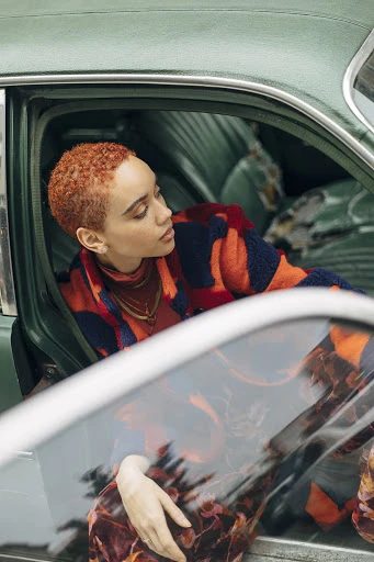 Stylish young woman with auburn hair poses inside a vintage turquoise car, wearing an orange and navy blue striped coat.