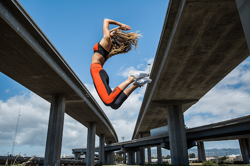 Athletic woman in sports bra and leggings jumps mid-air between concrete bridges against a blue sky with scattered clouds.