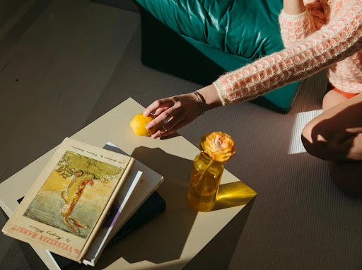 Woman reaches for lemon on table with books and flower in vase. Indoor setting with soft natural light and a cozy, domestic atmosphere.