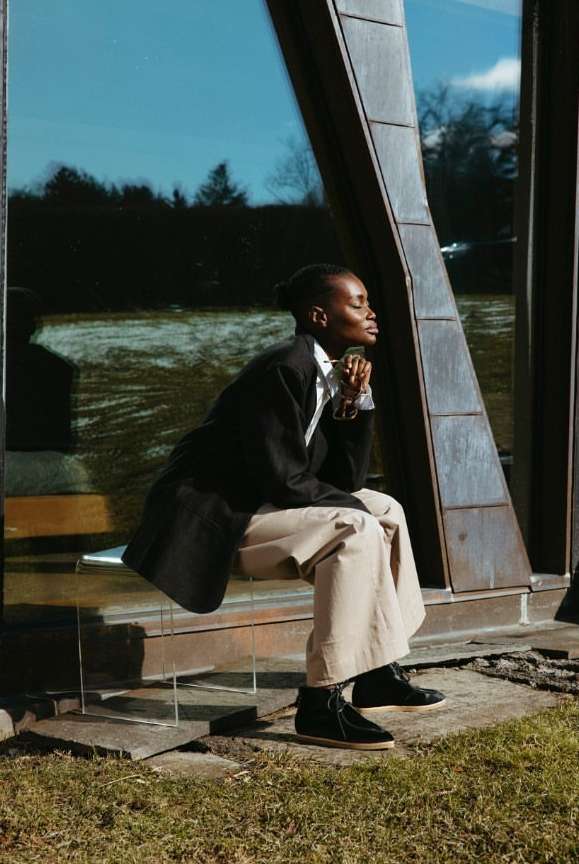 Woman in black coat and beige pants seated on transparent bench near modern building with blue sky reflection, outdoor.