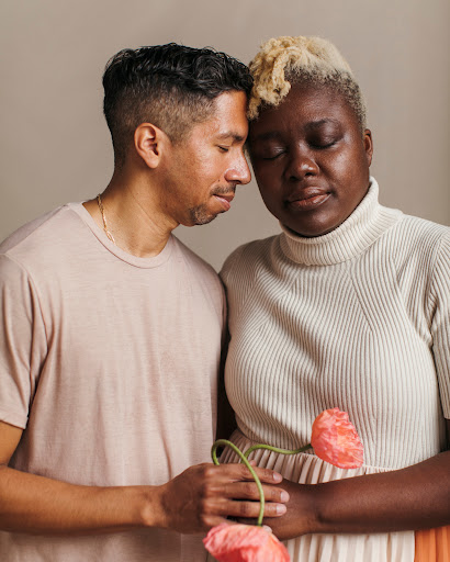 Portrait of a mixed-race couple with closed eyes. The woman has blonde hair and holds two pink flowers in her hand. Indoor setting.
