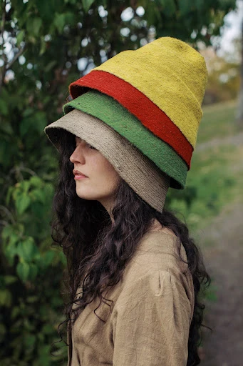 Woman in neutral clothing wearing a stack of three layered hats with yellow, red, green, and neutral colors in an outdoor setting.