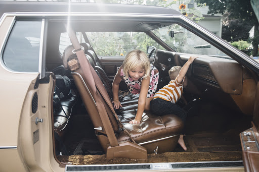 Two children, a girl and a boy, are sitting inside of an antique car with brown leather interior, and a tan exterior parked outside.
