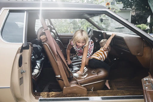 Two children, a girl and a boy, are sitting inside of an antique car with brown leather interior, and a tan exterior parked outside.