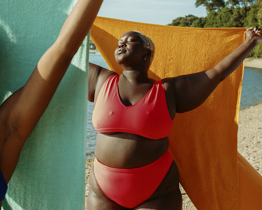Woman in a red bikini holds a towel on a sunny beach with water and trees in the background, enjoying summer on the beach.