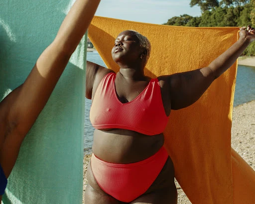 Woman in a red bikini holds a towel on a sunny beach with water and trees in the background, enjoying summer on the beach.