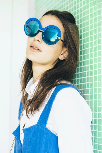 Brunette woman in blue sunglasses and overalls leans against a green tiled wall in a fashion portrait with a soft, diffused light.
