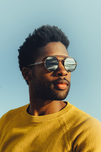 Close-up of a stylish African American man wearing round sunglasses and a yellow sweater against a clear blue sky backdrop.
