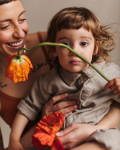 A mother and child pose indoors with orange flowers, with the mother smiling and the child looking at the camera with a serious expression.