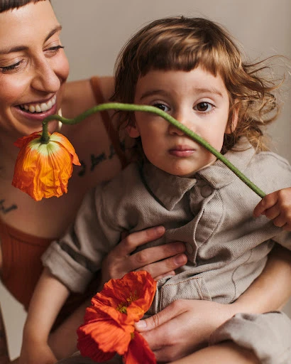 A mother and child pose indoors with orange flowers, with the mother smiling and the child looking at the camera with a serious expression.