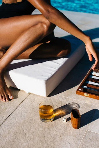 Woman relaxes poolside with backgammon game and a drink. Sunlit patio scene with leisure and recreation elements.