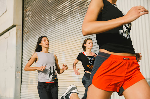 Three adult women are running outdoors on a city street, wearing athletic apparel on a sunny day in a medium eye-level shot.