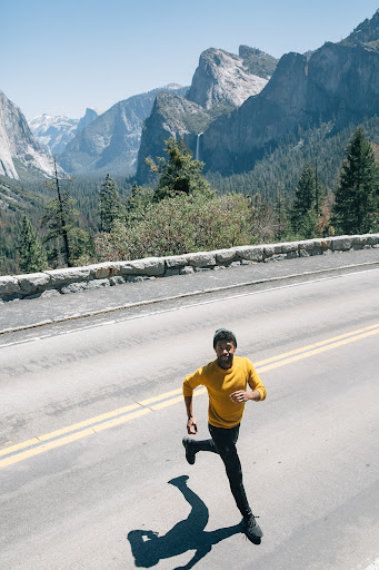 Athletic Black man in yellow shirt running on asphalt road with mountain landscape background on sunny day, fitness and outdoor activity.