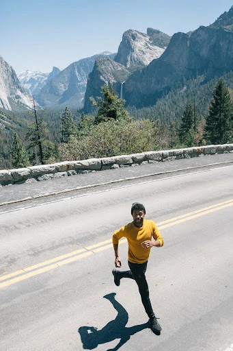 Athletic Black man in yellow shirt running on asphalt road with mountain landscape background on sunny day, fitness and outdoor activity.