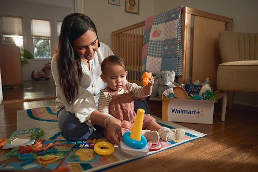 Mother and infant child play indoors with colorful toys on a rug, with a baby crib and a box of toys in the background.